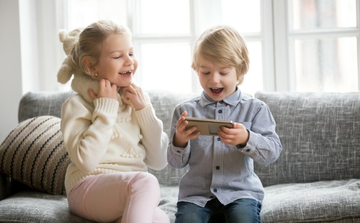 Excited kids having fun using smartphone sitting together on sof