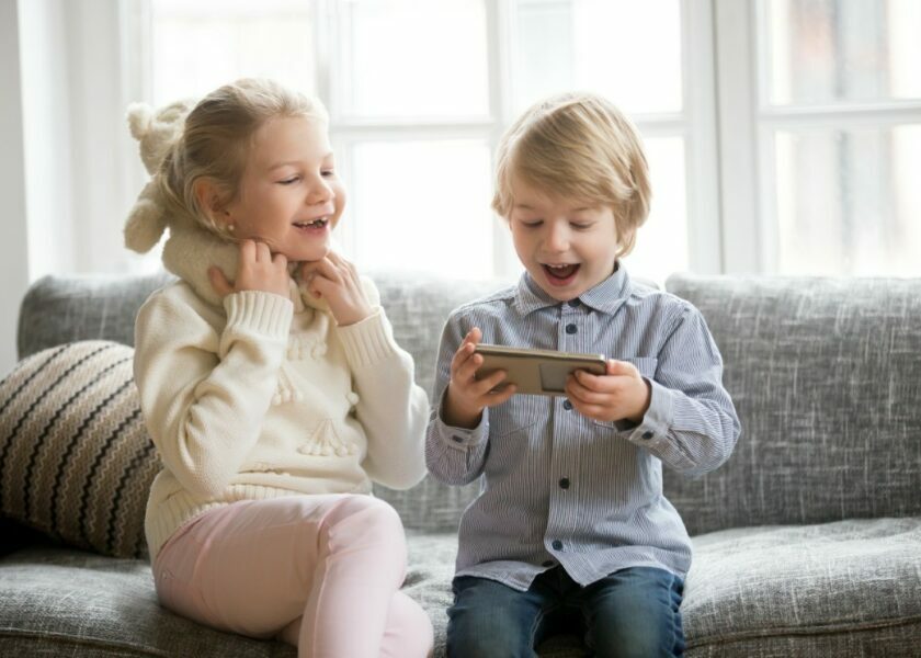 Excited kids having fun using smartphone sitting together on sof