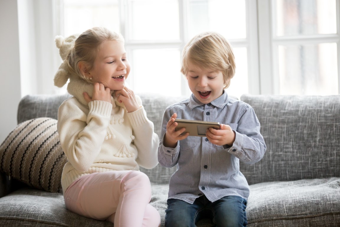 Excited kids having fun using smartphone sitting together on sof